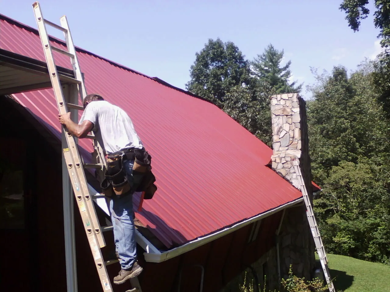 Roofer on a ladder beside a red metal roof and stone chimney — Soffit & Fascia Repair by Roofing Brothers in Gold Beach
